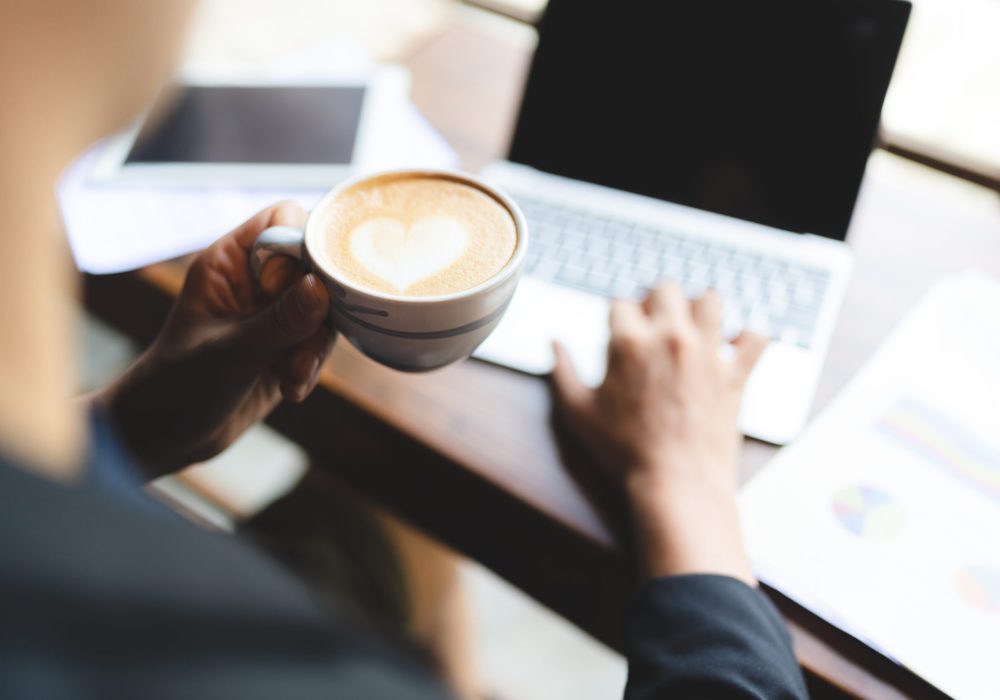 Rear closeup view of young business man and entrepreneur drinking drinking coffee with heart cream frosting and working on laptop in modern office and cafeteria